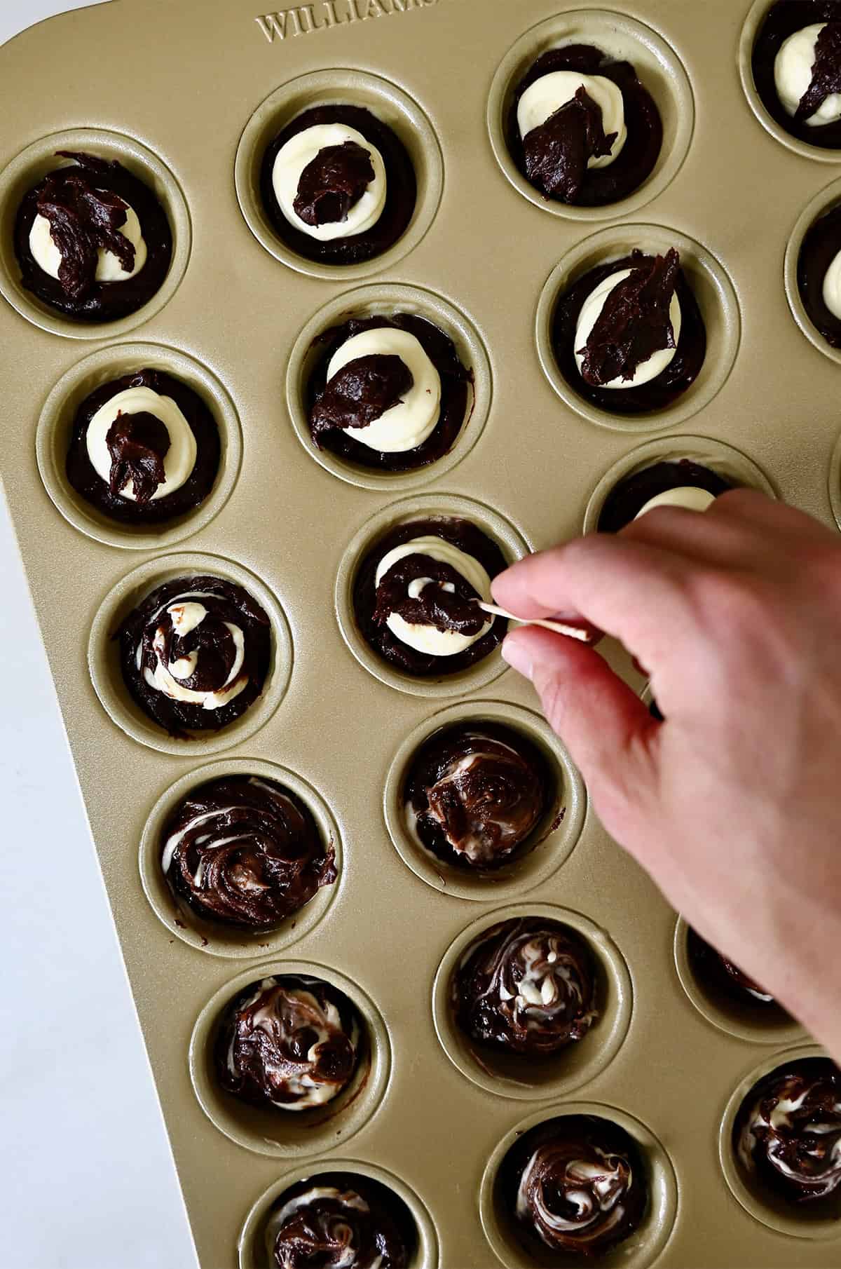 Cheesecake filling and brownie batter is swirled together with a toothpick in a mini muffin pan.