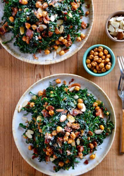 Rustic white plates hold a kale salad topped with sliced almonds and crispy chickpeas. Nearby are small bowls of crispy chickpeas and sliced almonds.