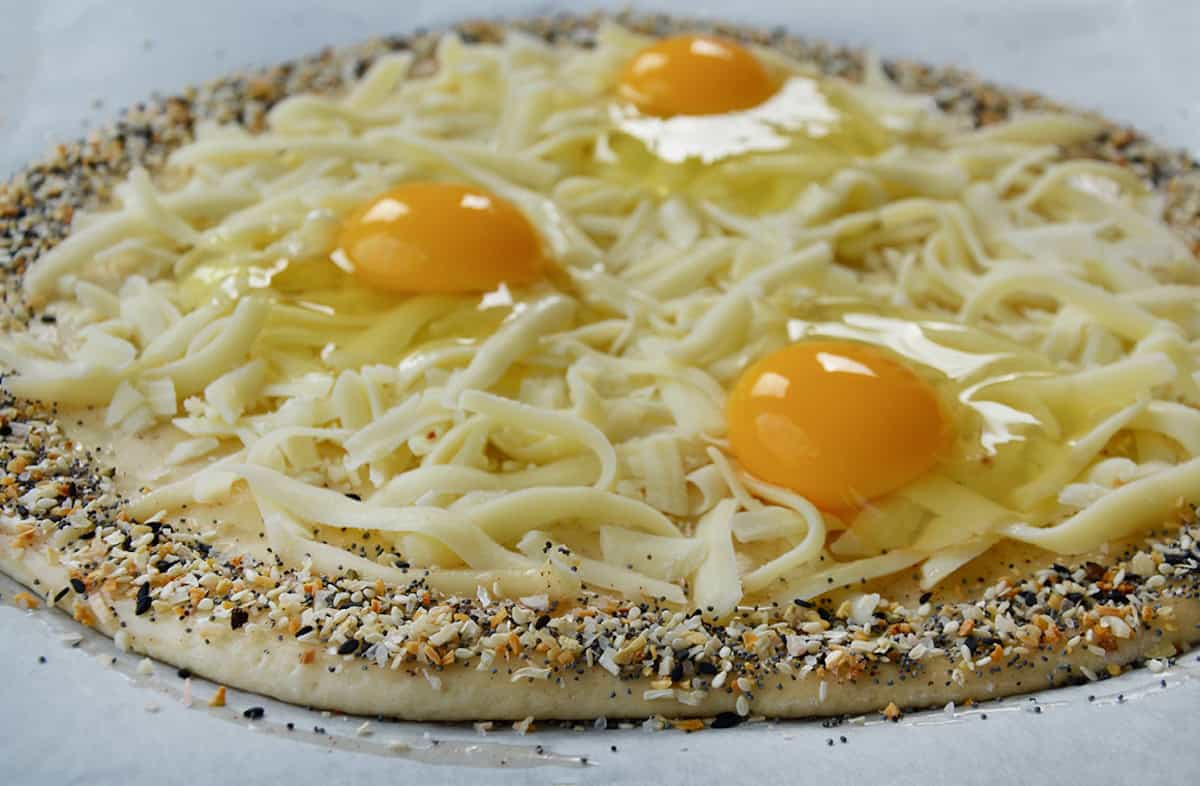 Pizza dough sprinkled with everything bagel seasoning and topped with grated mozzarella and three raw eggs. 