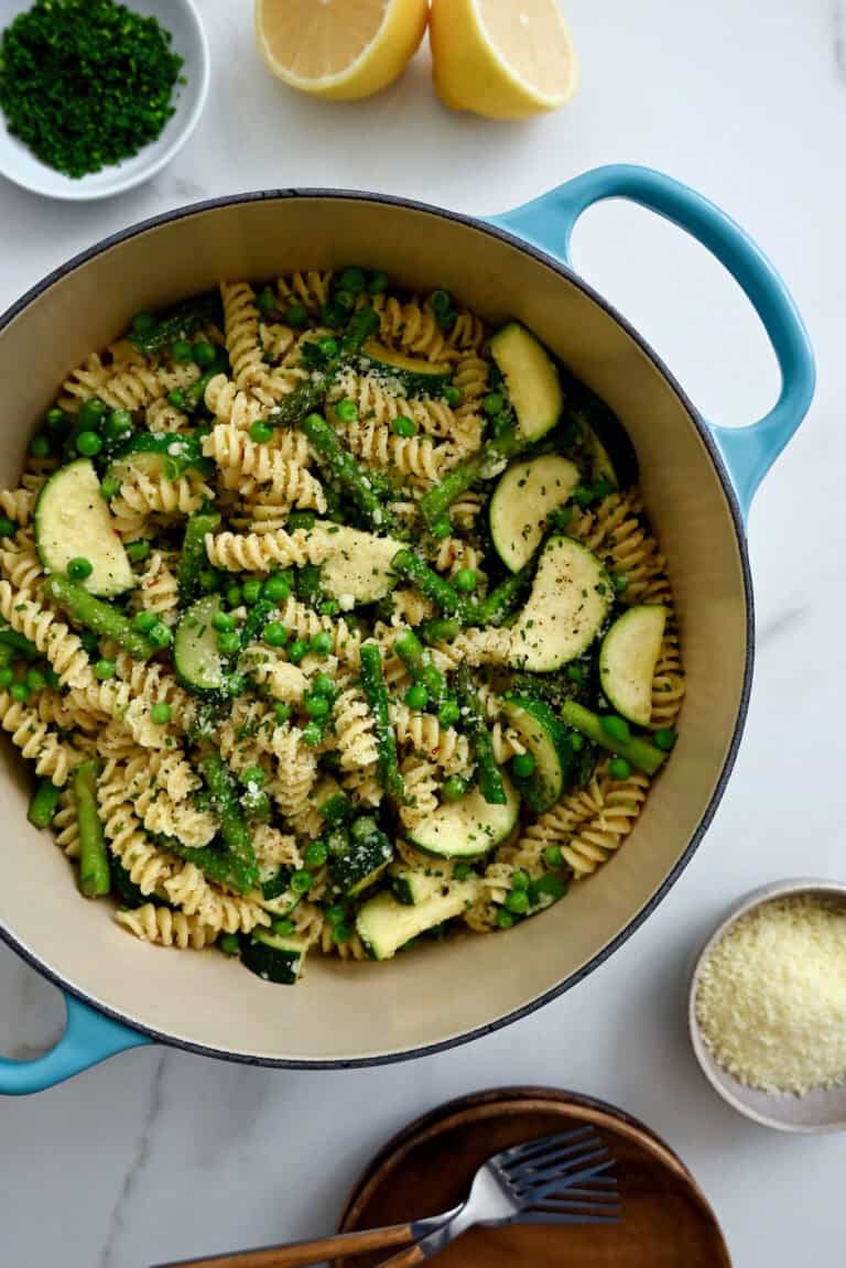 A blue Dutch oven is filled with pasta primavera made with peas, asparagus and zucchini. A little dish of parmesan and a stack of rustic bowls and forks sit nearby.