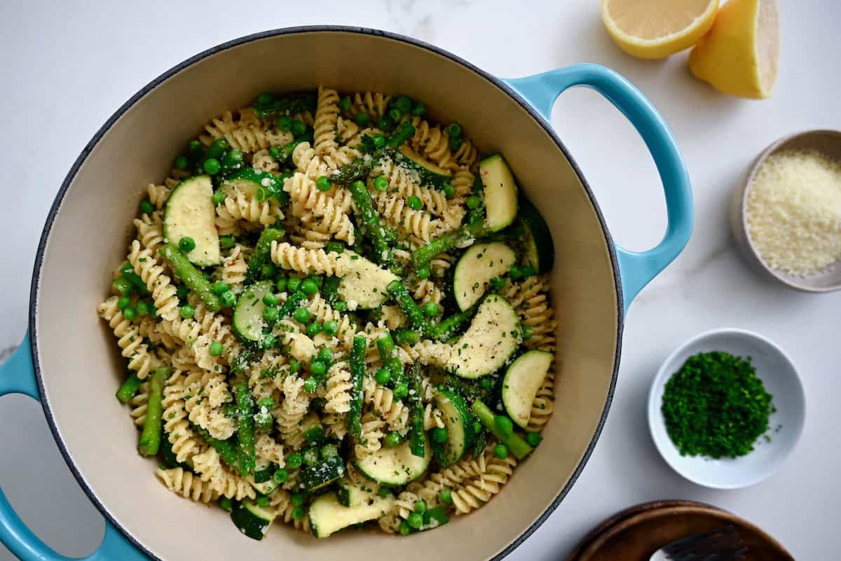 A blue Dutch oven is filled with pasta primavera made with peas, asparagus and zucchini. Dishes of parmesan and minced chives, lemon halves and a partially visible stack of rustic bowls and forks sit nearby.