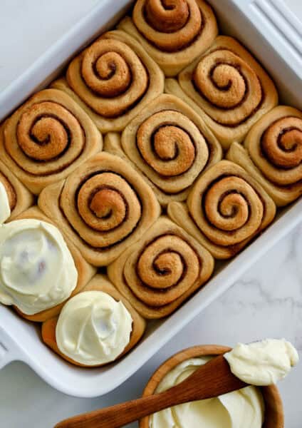 Cinnamon rolls in a white baking dish are partially frosted with cream cheese frosting. A wooden bowl with cream cheese frosting and a spatula is nearby.