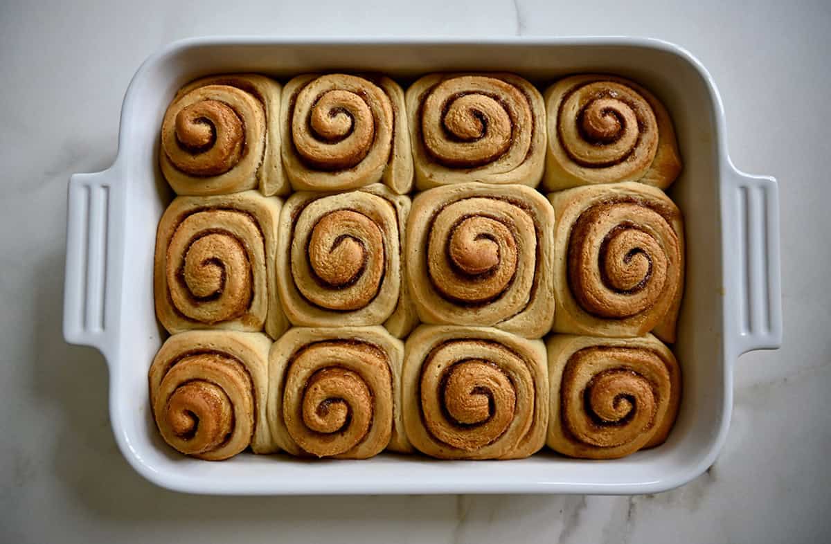 A white rectangular baking dish holds a dozen baked and unfrosted cinnamon rolls.