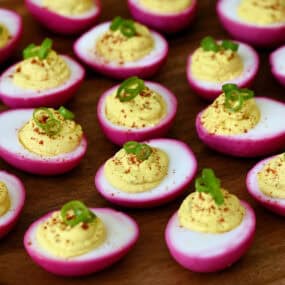 Pickled deviled eggs, naturally dyed pink with beets and garnished with paprika and scallions, are arranged on a round wooden board.