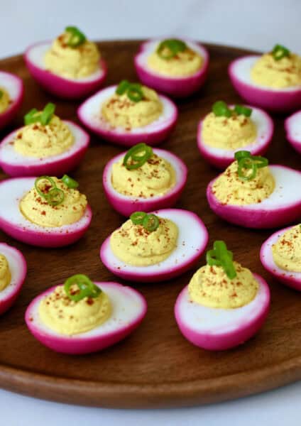 Pickled deviled eggs, naturally dyed pink with beets and garnished with paprika and scallions, are arranged on a round wooden board.