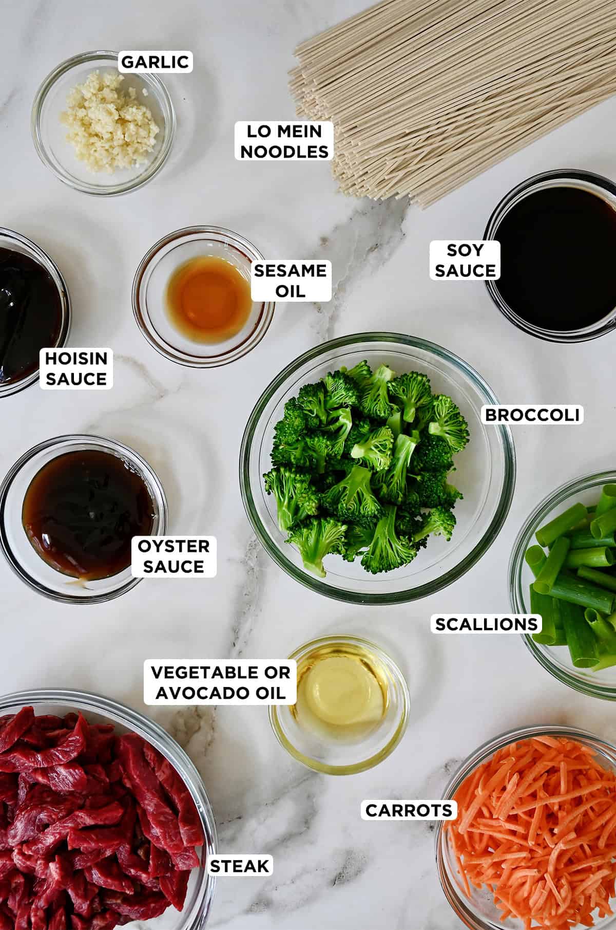 Various sizes of glass bowls containing beef lo mein ingredients, including flank steak pieces, oyster sauce, hoisin sauce, sesame oil, broccoli florets, vegetable oil, scallions, shredded carrots, soy sauce, lo mein noodles and minced garlic.