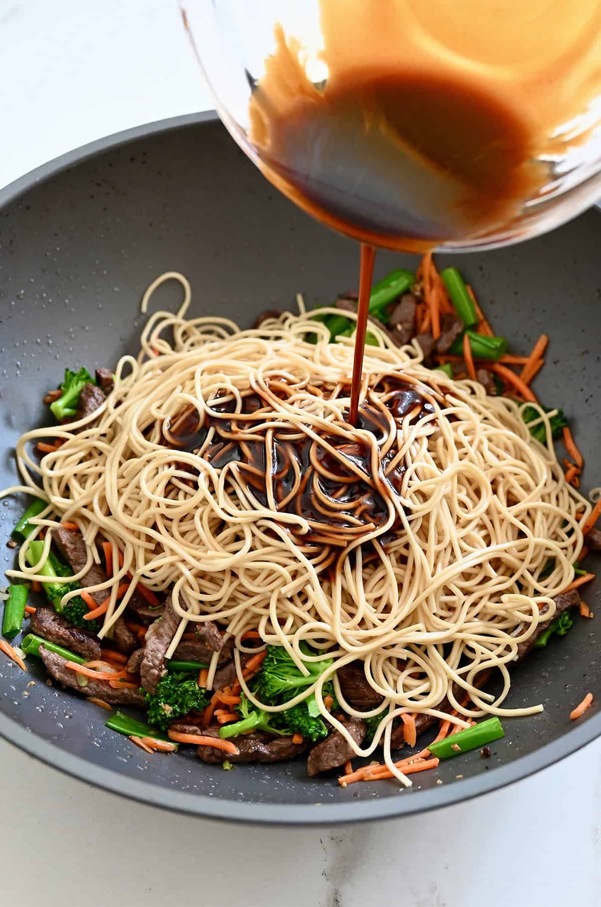 Lo mein sauce being poured over noodles and stir-fried veggies in a wok.