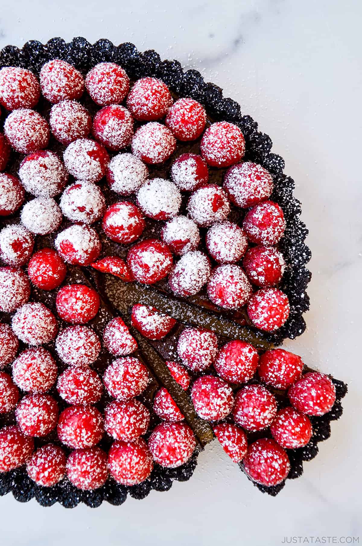 A chocolate tart with fresh raspberries on top and dusted with powdered sugar. A piece is cut out of the tart.