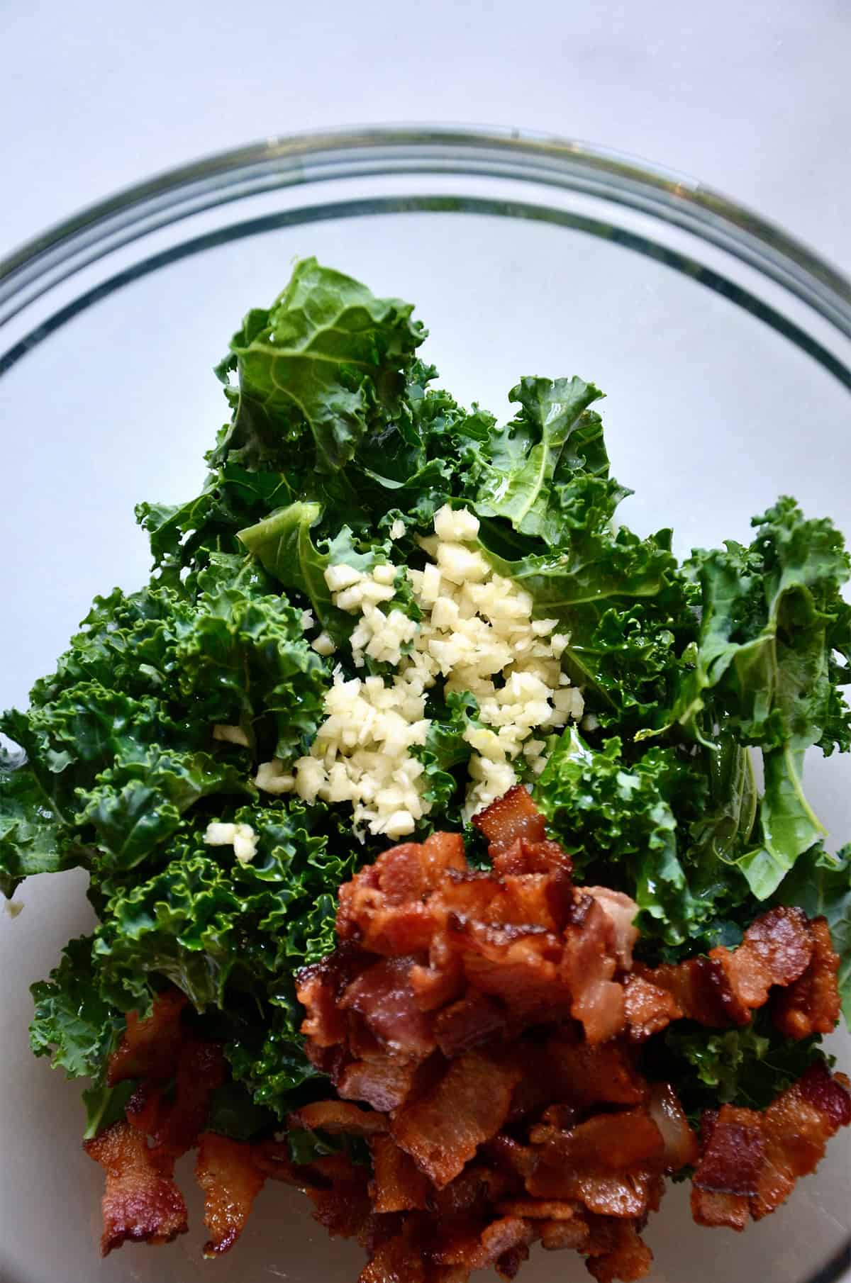Chopped kale, crispy pieces of bacon and minced garlic in a large glass bowl.