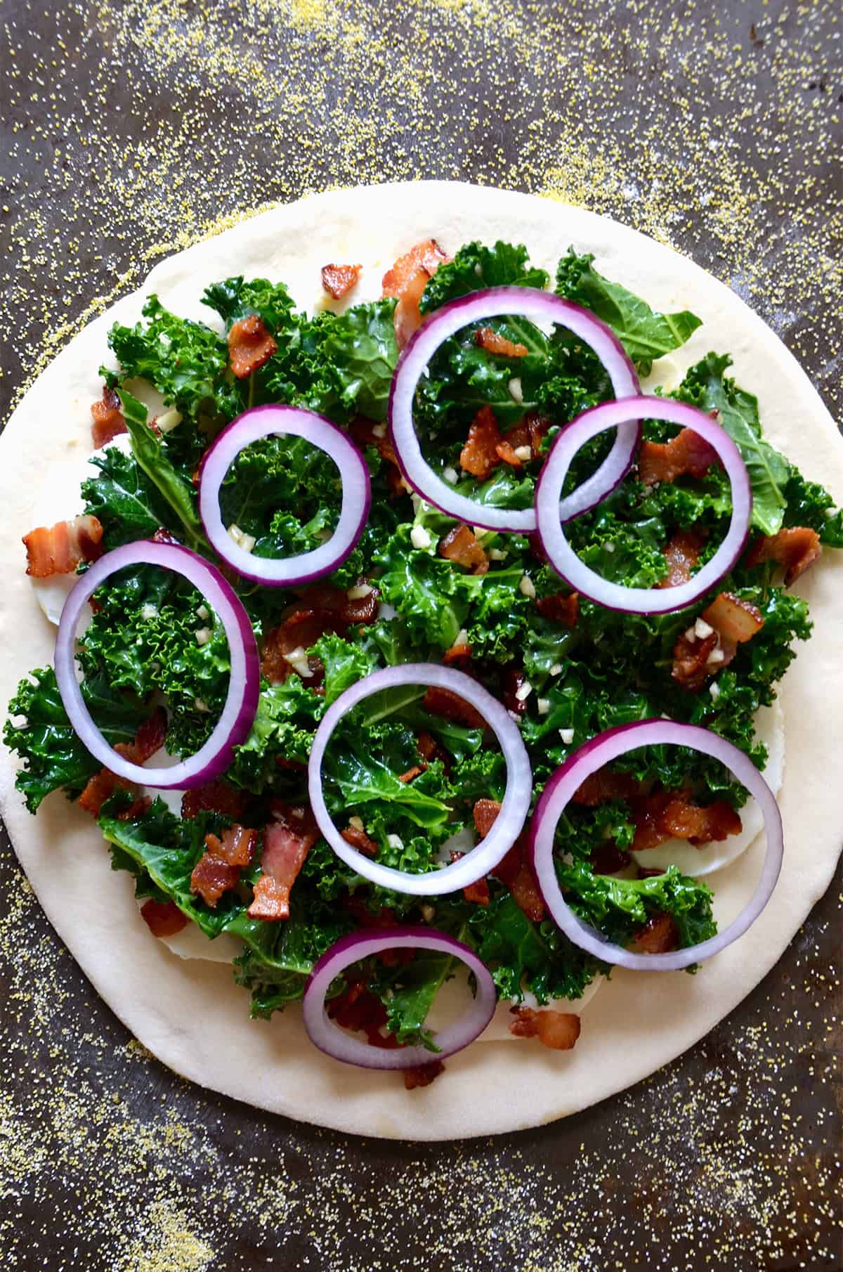 A round of pizza dough topped with kale, chopped, crispy bacon and sliced red onion on a cornmeal-dusted surface.