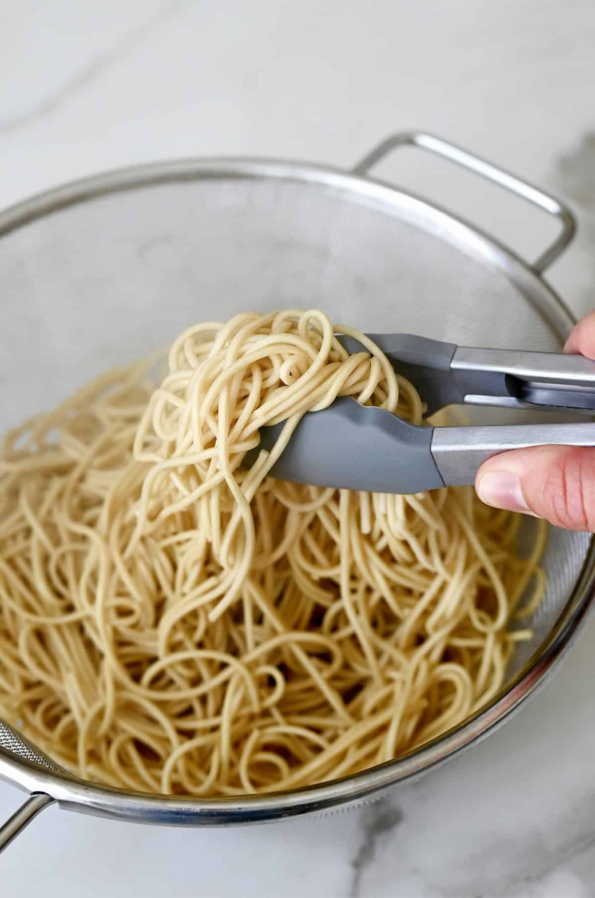 Tongs grabbing cooked lo mein noodles from a mesh strainer.