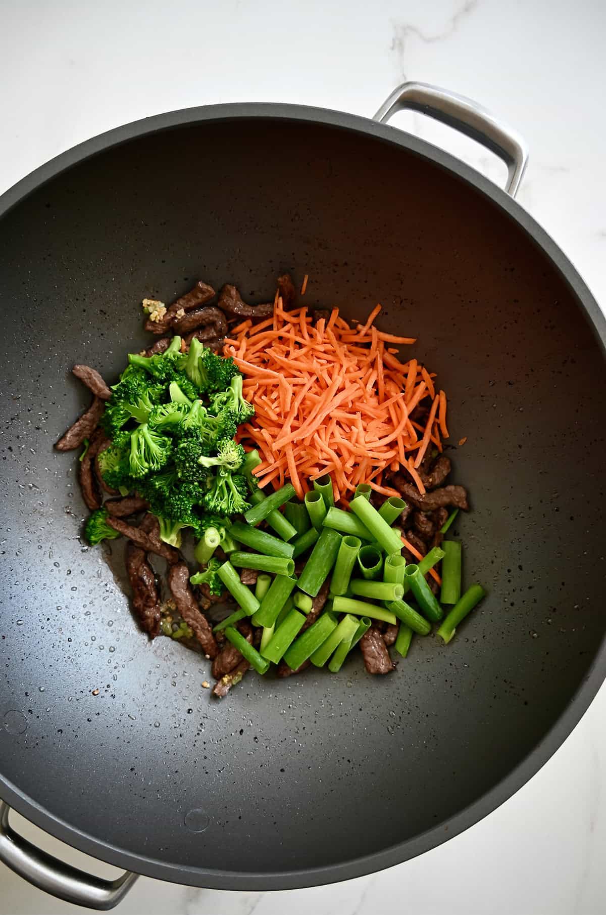 A wok with cooked beef strips, shredded carrots, broccoli and sliced scallions.