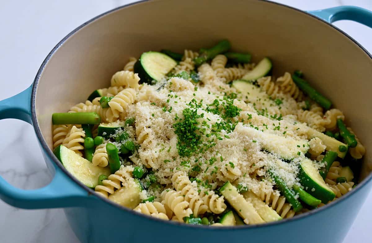 A Dutch oven is filled with pasta tosswed with zucchini, asparagus and peas. The mixture is sprinkled generously with parmesan cheese and minced chives.