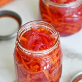 Two glass jars are filled with pickled red onions and carrots. The lids and some carrots sit nearby.