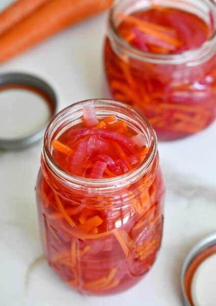 Two glass jars are filled with pickled red onions and carrots. The lids and some carrots sit nearby.