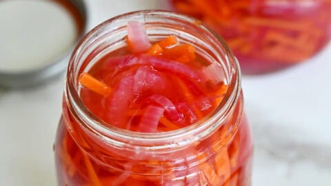 Two glass jars are filled with pickled red onions and carrots. The lids and some carrots sit nearby.