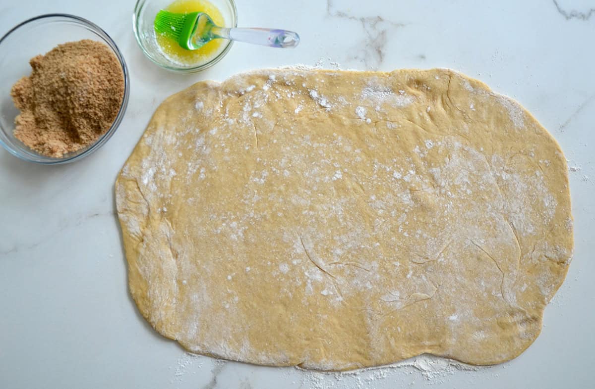 Cinnamon roll dough is rolled out on a white marble work surface. Glass bowls containing melted butter and a cinnamon sugar mixture are nearby.