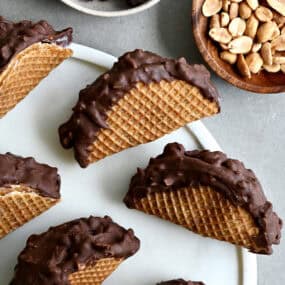 Homemade choco tacos on a round serving tray next to a bowl containing chocolate chips and another bowl containing peanuts.