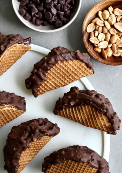 Homemade choco tacos on a round serving tray next to a bowl containing chocolate chips and another bowl containing peanuts.