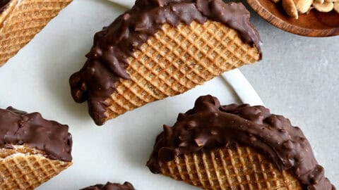 Homemade choco tacos on a round serving tray next to a bowl containing chocolate chips and another bowl containing peanuts.