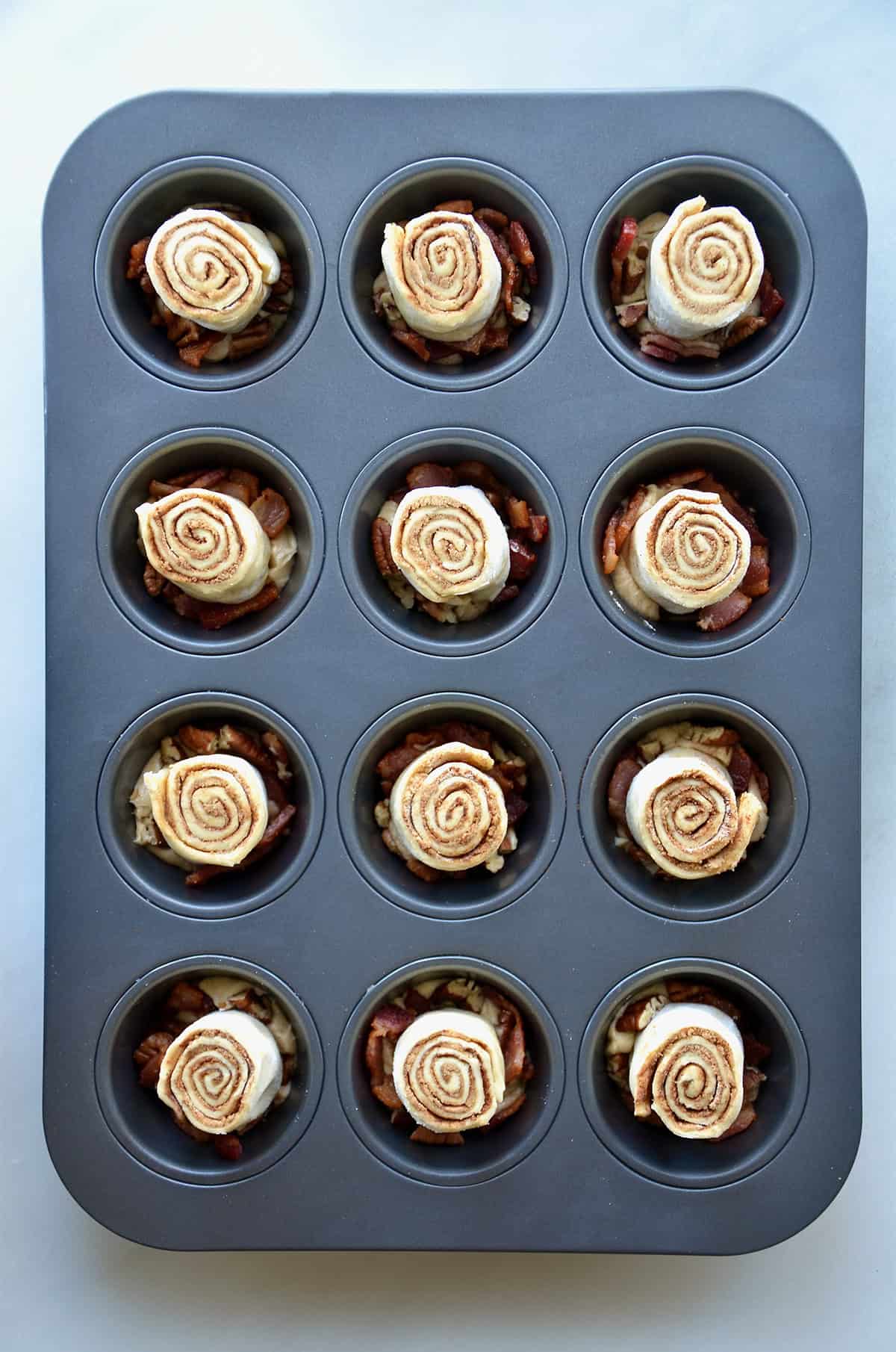 Portions of cinnamon rolls on top of mounds of butter and chopped bacon and pecans in the wells of a muffin tin.