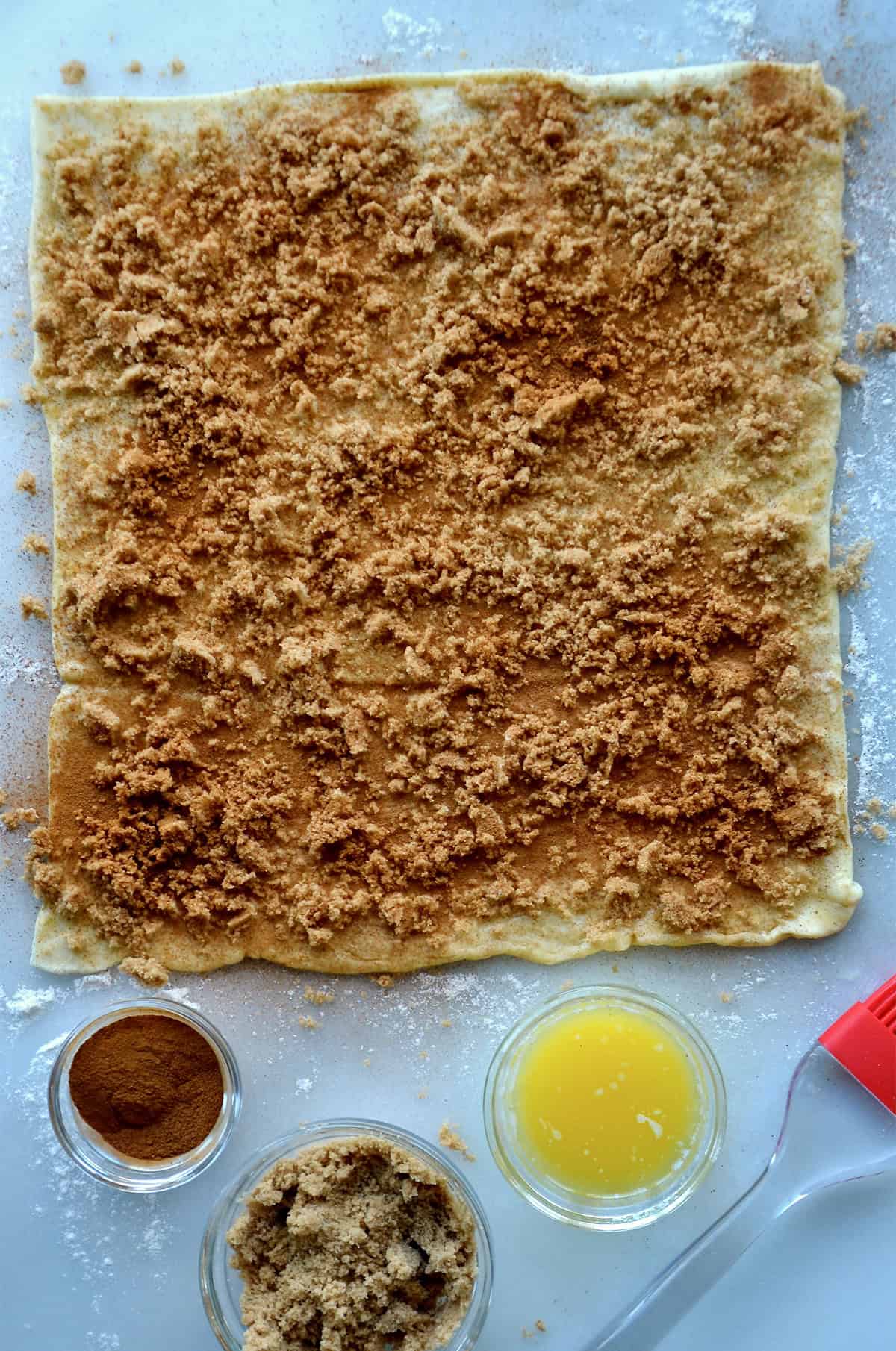 A rectangle of puff pastry topped with melted butter, brown sugar and ground cinnamon. Small bowls of melted butter, brown sugar and cinnamon are beside the dough.