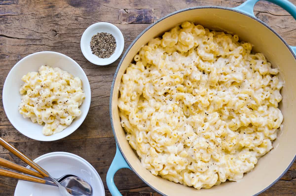 A blue Dutch oven filled with white cheddar mac and cheese sits on a wooden surface. A portion sits nearby in a white bowl, next to a stack of empty bowls and spoons. A small white bowl containing black pepper is also visible.