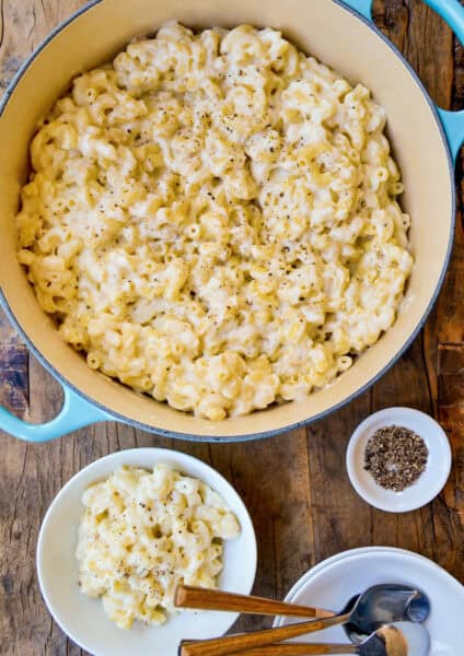A blue Dutch oven filled with white cheddar mac and cheese sits on a wooden surface. A portion sits nearby in a white bowl, next to a stack of empty bowls and spoons. A small white bowl containing black pepper is also visible.
