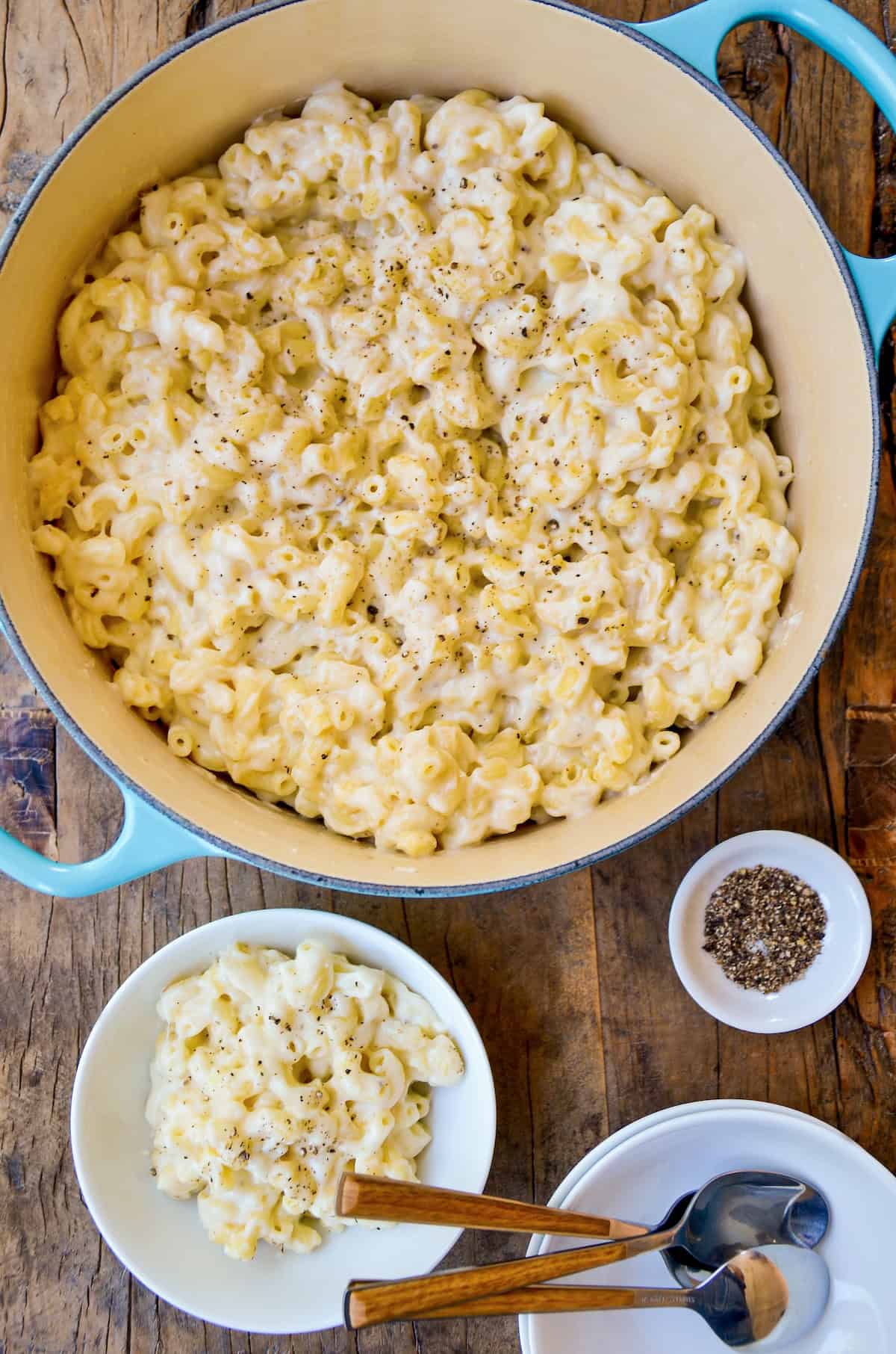 A blue Dutch oven filled with white cheddar mac and cheese sits on a wooden surface. A portion sits nearby in a white bowl, next to a stack of empty bowls and spoons. A small white bowl containing black pepper is also visible.