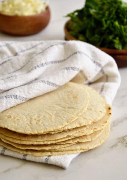 A stack of homemade corn tortillas are tucked into a striped dishtowel. They sit on a marble counter, with wooden bowls containing grated cheese and chopped herbs in the background.