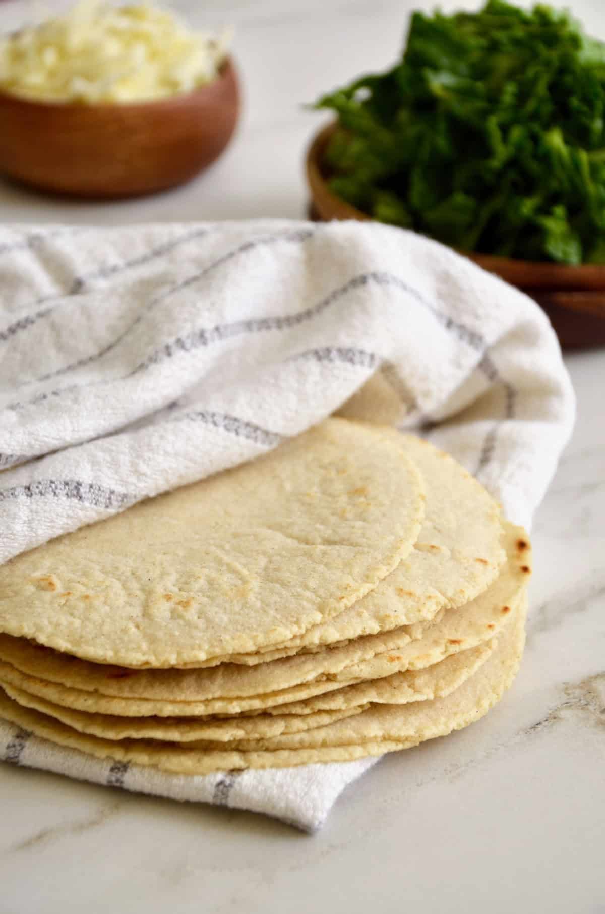 A stack of homemade corn tortillas are tucked into a striped dishtowel. They sit on a marble counter, with wooden bowls containing grated cheese and chopped herbs in the background.