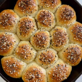 Golden brown bagel bites sprinkled with everything seasoning are arranged in a cast iron skillet on a white marble surface.