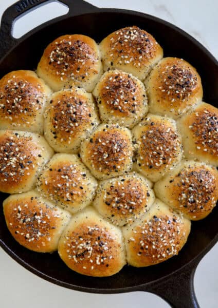 Golden brown bagel bites sprinkled with everything seasoning are arranged in a cast iron skillet on a white marble surface.
