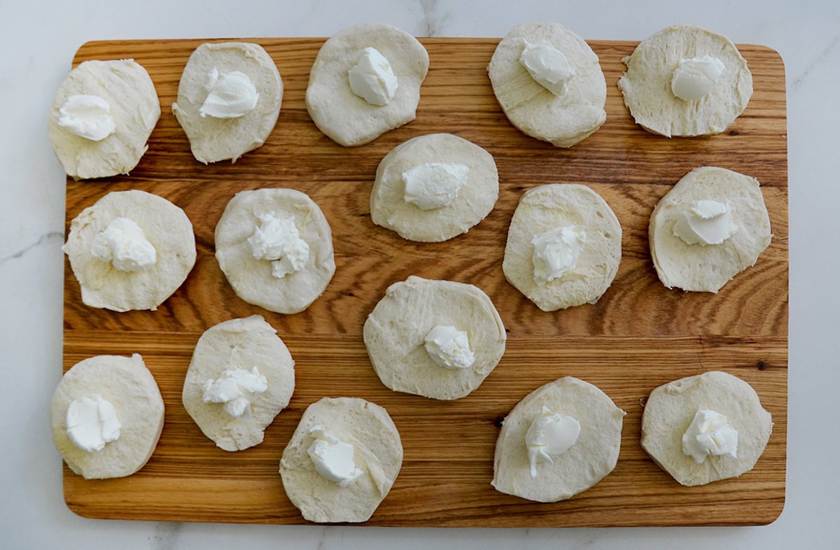 Round pieces of biscuit dough are topped with dollops of cream cheese. They're arranged on a wooden board on a white marble surface.