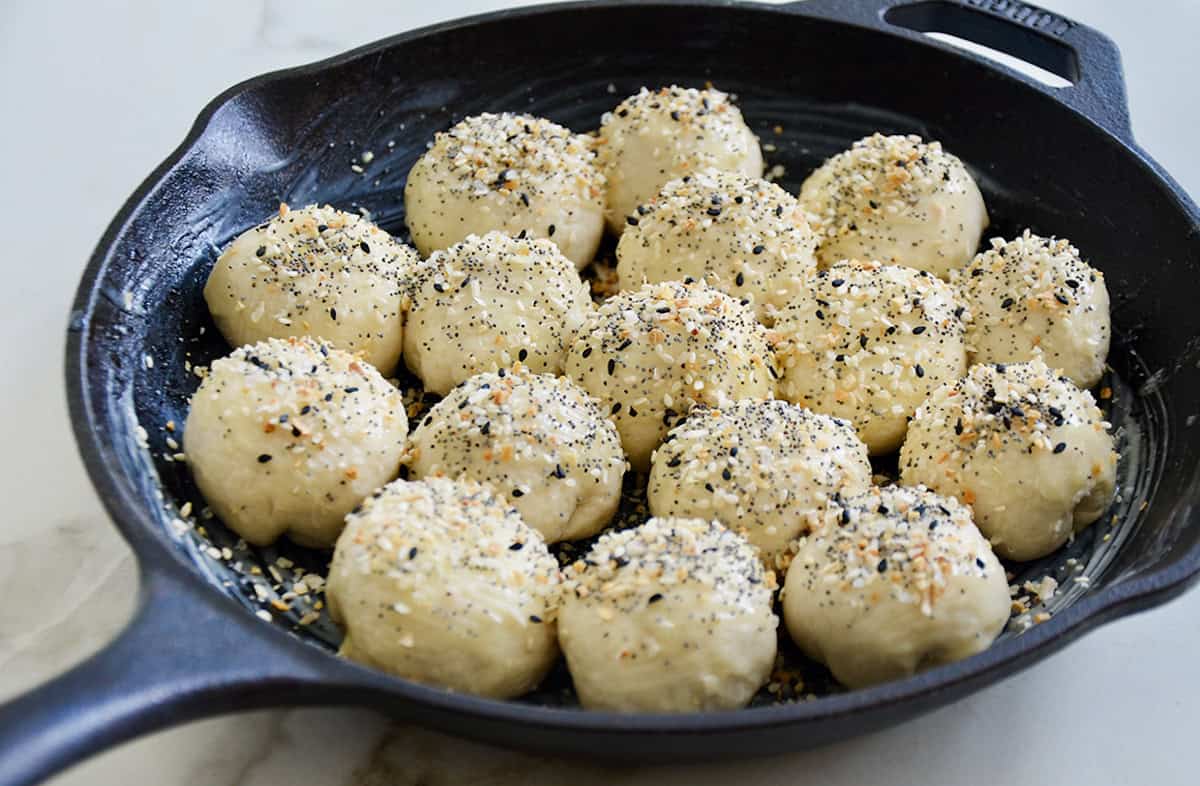 Unbaked bagel bites sprinkled with everything seasoning are arranged in a cast iron skillet.