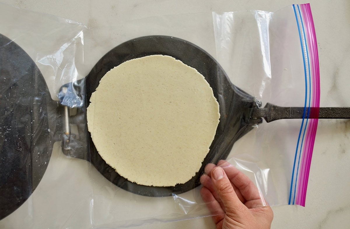 A corn tortilla is formed with a black cast iron tortilla press. A cut-open plastic resealable bag lines the press.