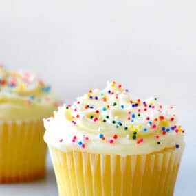 A vanilla cupcake is topped with vanilla buttercream and rainbow sprinkles. It sits on a white surface with another cupcake in the background.