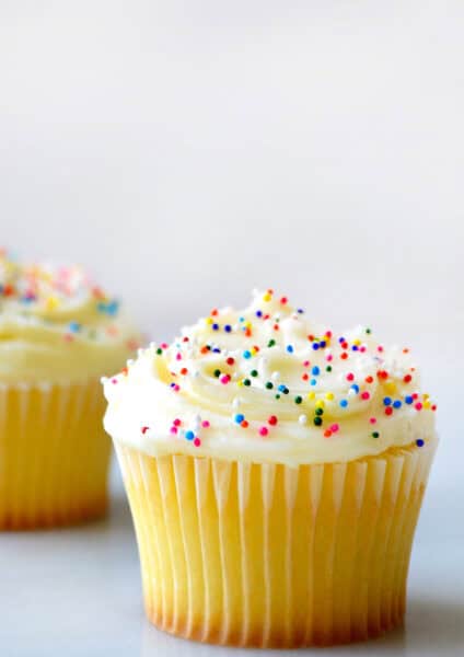 A vanilla cupcake is topped with vanilla buttercream and rainbow sprinkles. It sits on a white surface with another cupcake in the background.