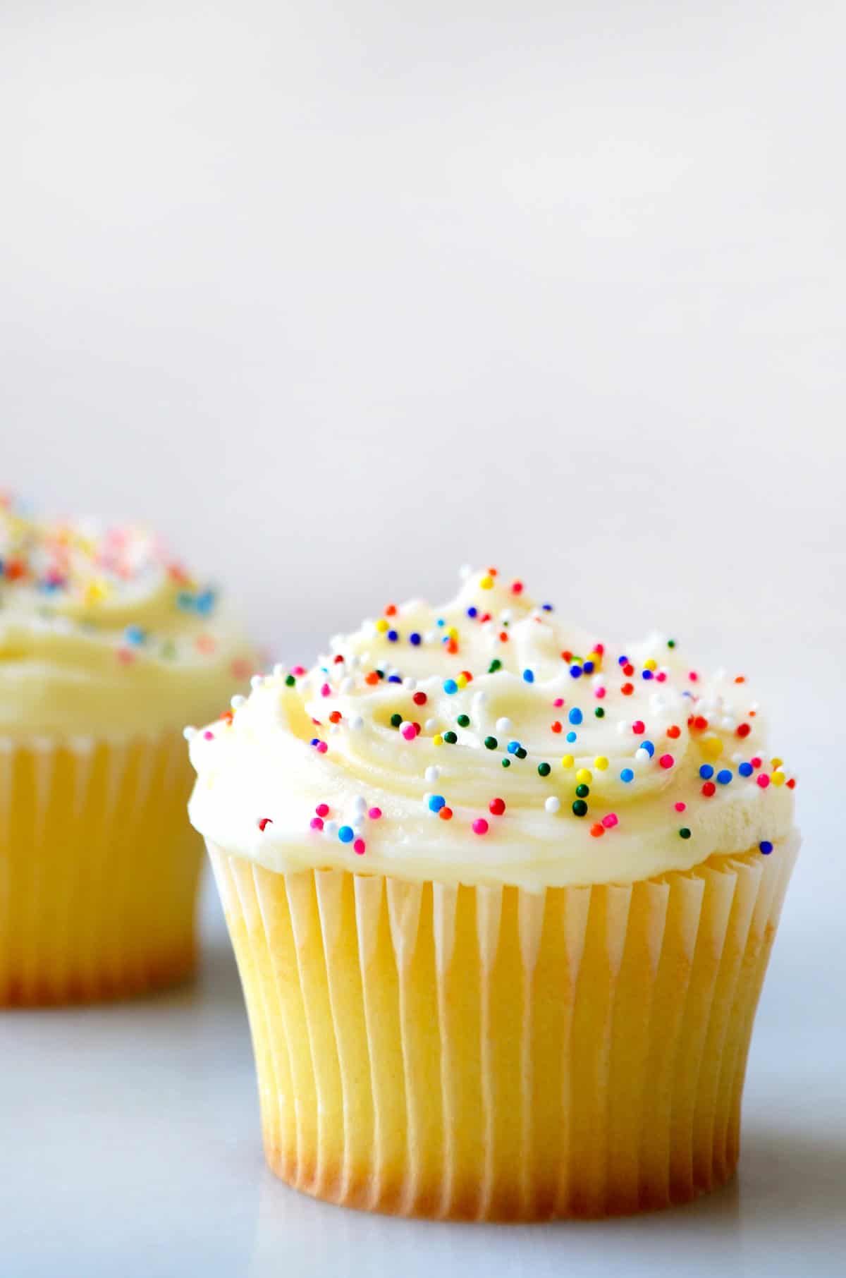 A vanilla cupcake is topped with vanilla buttercream and rainbow sprinkles. It sits on a white surface with another cupcake in the background.