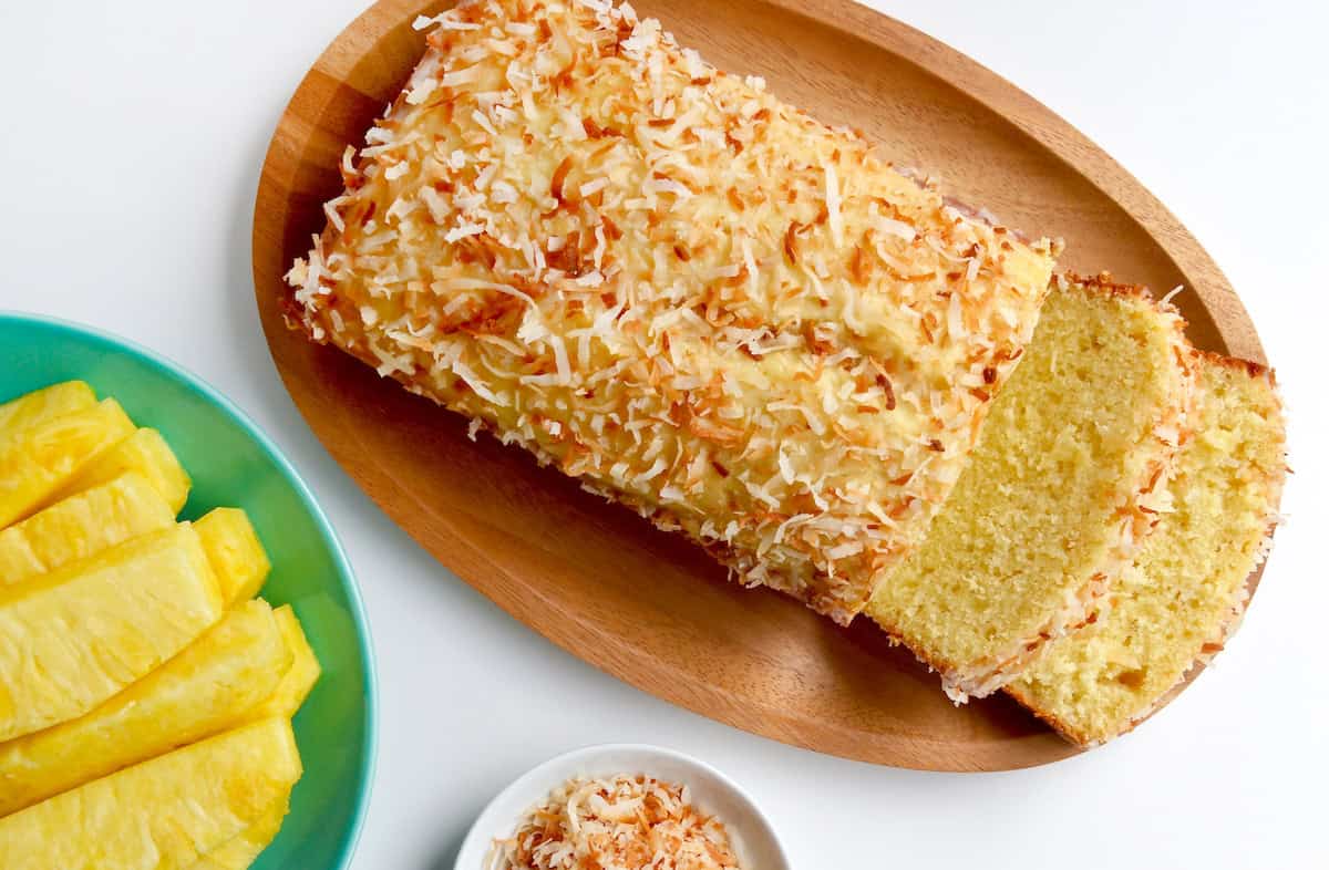 A pineapple coconut loaf with two slices cut off of it. Slices of pineapple and a bowl of toasted coconut are beside the loaf.