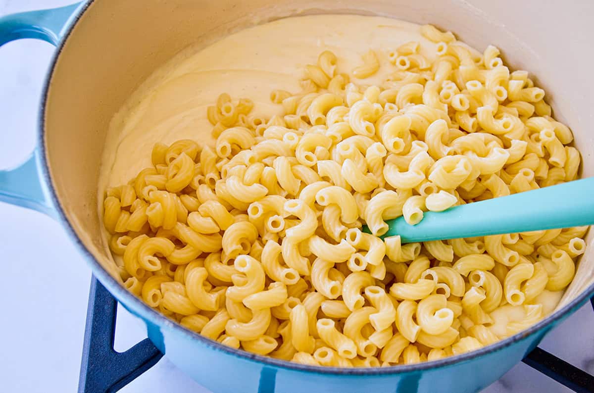 In a blue Dutch oven, a blue spoon stirs cooked cavatappi pasta into a creamy cheese sauce.