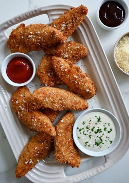 Crispy chicken tenders topped with grated parmesan cheese on a serving platter with small bowls of dipping sauces.