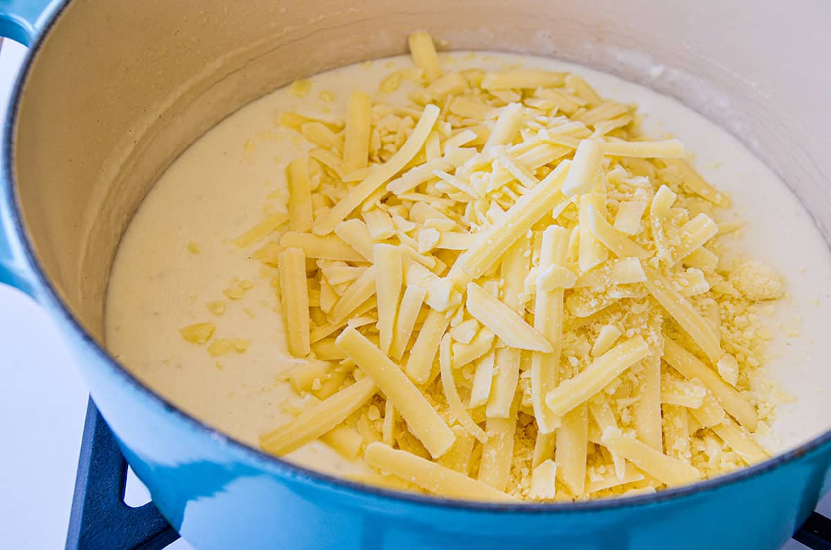 In a blue Dutch oven, grated white cheddar and parmesan cheese is sprinkled onto cream sauce.