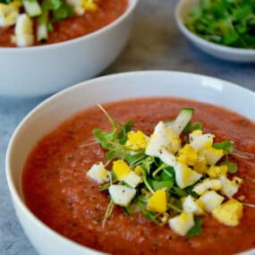A white bowl of gazpacho is garnished with chopped cooked egg and microgreens. In the background is another filled bowl of gazpacho and small dishes of the chopped egg and greens.