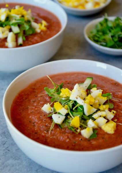 A white bowl of gazpacho is garnished with chopped cooked egg and microgreens. In the background is another filled bowl of gazpacho and small dishes of the chopped egg and greens.