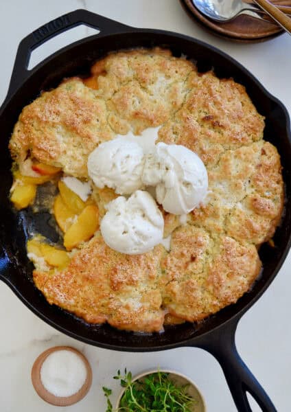 Peach cobbler with buttermillk biscuits is topped with melting vanilla ice cream in a cast iron skillet.Little dishes containing sanding sugar and fresh thyme are nearby.