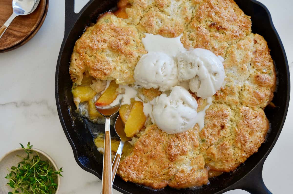 Peach cobbler with buttermillk biscuits is topped with melting vanilla ice cream in a cast iron skillet. Two wood-handled spoons stick out of the pan and a little dish with fresh thyme is nearby.