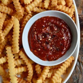 A white dish of spicy ketchup sits in a big bowl of crinkle-cut French fries.