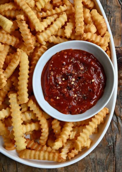 A white dish of spicy ketchup sits in a big bowl of crinkle-cut French fries.