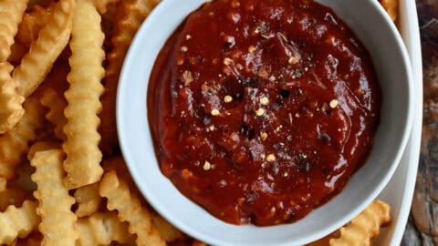 A white dish of spicy ketchup sits in a big bowl of crinkle-cut French fries.